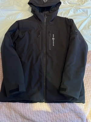 Svart Sail Racing-jacka – Mycket bra skick - Snygg och funktionell svart jacka från Sail Racing i storlek 160. Jackan är i fint skick och perfekt för både vardag och utomhusaktiviteter. Den är tillverkad i ett slitstarkt, vind- och vattenavvisande material som ger bra skydd i varierande väder.  Detaljer: 	•	Färg: Svart 	•	Storlek: 160 	•	Justerbar och skyddande huva 	•	Full längd-dragkedja 	• Två	bröstfickor med vattentät dragkedja 	•	Två rymliga sidfickor 	•	Sail Racing-logga på bröstet 	•	Lätt och bekväm passform