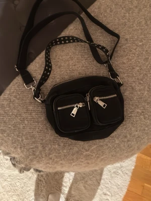 Svart axelväska med nitar och dragkedjor - Cool svart axelväska i tyg med två framfickor med silvriga dragkedjor och en bred axelrem dekorerad med nitar. Väskan har justerbar rem och detaljer i metall, perfekt för dig som gillar en edgy stil.