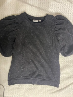 Glittrig topp - Supersnygg svart topp från Kappahl med glittrigt tyg och stora puffärmar. Toppen har en rak passform och är perfekt för dig som vill sticka ut lite extra. Materialet känns mjukt och stretchigt, vilket gör den bekväm att bära. Enkel rund halsringning.