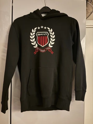Svart hoodie med GANT-logga - Snygg svart hoodie från GANT med stor broderad logga. Materialet är mjukt och bekvämt, perfekt för en chill och sportig stil. 