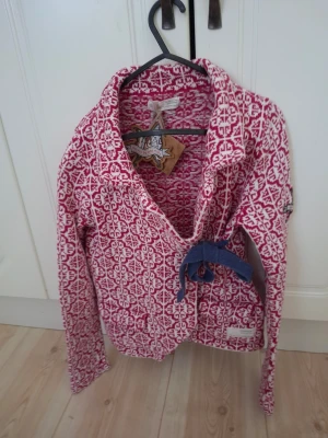 Rosa mönstrad kofta Odd Molly - Stickad kofta från Odd Molly i rosa och vitt med ett snyggt grafiskt mönster. Koftan har omlottmodell med blått knytband i midjan och en liten etikett nertill. Perfekt för dig som gillar färg och unika detaljer. Ny med prislapp 