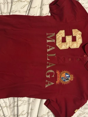Röd polotopp Malaga med brodyr - Snygg röd polotopp med stora broderade detaljer, texten 'MALAGA' och siffran 3 i beige på både fram- och baksida. Märkesemblem med sköld och krans på bröstet. Klassisk pikékrage och knappar framtill. Perfekt för en sportig och trendig look.