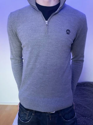 Grå halvzip tröja från J.Lindeberg - Snygg grå stickad tröja från J.Lindeberg med halv dragkedja och hög krage. Tröjan har diskret logga på bröstet, ribbade muddar och är tillverkad i mjukt material som passar perfekt till jeans eller chinos.