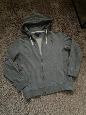 Grå zip-up hoodie från US Polo Assn - Grå hoodie med dragkedja från US Polo Assn. Klassisk modell med huva och vita snören! fraktar inom 24h📦 köp mer än ett plagg för 20% rabatt🤝 tveka inte på att höra av er vid frågor eller funderingar!
