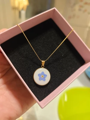 Guldhalsband med förgätmigej - Snyggt halsband med tunn guldfärgad kedja och runt hänge i vitt med en förgätmigej i mitten. Perfekt för dig som gillar unika och söta accessoarer. Rostfritt stål.