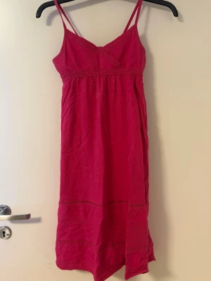 Rosa kort klänning med spetsdetaljer - Söt rosa kortklänning med tunna axelband och spetsdetaljer vid byst och nederkant. Klänningen har en v-ringning och är tillverkad i ett mjukt bomullstyg som passar perfekt till varma dagar.