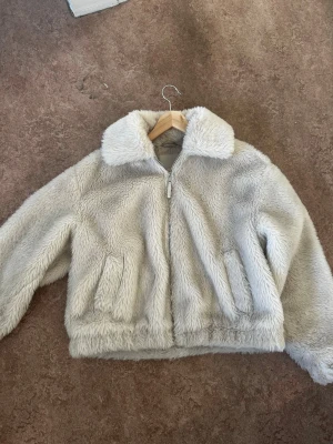 Beige päls jacka med dragkedja - Croppad beige  päls/teddy jacka, den ser lite grå ut på bild men är inte alls det I verkligheten bara beige. Stora fickor varm och jätte skön perfekt för vår vinter och höst