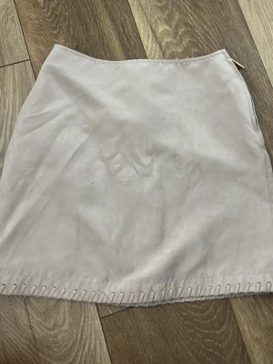 Beige mockakjol med dekorativ söm - Snygg beige kortkjol i mockaimitation med dekorativ söm längs nederkanten. Kjolen har en enkel, rak passform och dragkedja i sidan för enkel på- och avtagning. Perfekt att matcha med en stickad tröja eller t-shirt.