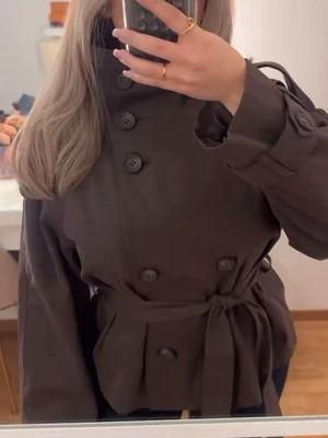 Kort brun trenchcoat från Bershka - Snygg brun trenchcoat med dubbelknäppning och höga knappar. Jackan är från Bershka och är i storlek M, den är slutsåld just nu så svårt att få tag på den.