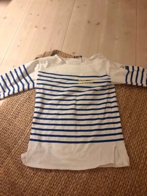 Randig vit och blå topp från Armor Lux - Säljer en klassisk vit topp från Armor Lux med blå horisontella ränder och trekvartsärm. Toppen har en liten slits i sidan och logotyp på ärmen. Tillverkad i mjuk bomull, perfekt för en avslappnad och stilren look.