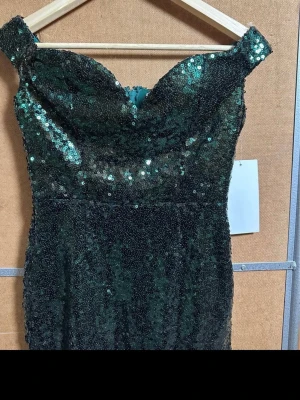 Grön paljettklänning offshoulder - Snygg grön kortklänning täckt av glittriga paljetter. Offshoulder-modell med en smickrande v-ringning och figurnära passform. Perfekt för dig som vill sticka ut med en glamorös look.