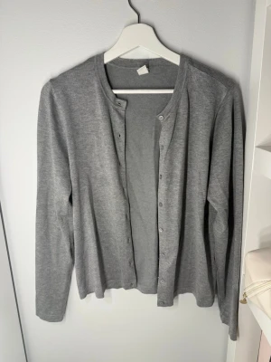 Grå basic kofta med rund hals - En stilren grå kofta med rund halsringning och knäppning framtill. Koftan har långa ärmar och är tillverkad i ett mjukt, tunt material som passar perfekt för lager-på-lager. Enkel och tidlös design som funkar till det mesta.