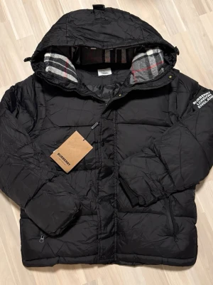 Svart pufferjacka från Burberry - Snygg svart pufferjacka från Burberry med klassiskt rutigt innerfoder i huvan och dragkedja framtill. Jackan har en patch med Burberry-logga på ärmen och praktiska fickor med dragkedja. Perfekt för kalla dagar och riktigt trendig vinterstil.