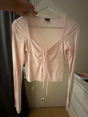Ljusrosa topp med snörning - Trendig ljusrosa topp med lång ärm och snygg snörning framtill. Djup v-ringning och mjukt material som känns skönt mot huden. Perfekt för dig som gillar en feminin och modern stil.