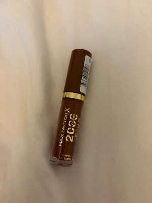 Max Factor Lipgloss Chocolate Glow - Max Factor X 2000 Calorie Lip Glaze i färgen 180 Chocolate Glow. Ett plumping läppglans i en snygg brun nyans med guldig detalj på hylsan. Ger glansiga och fylliga läppar med en hint av chokladton. Perfekt för dig som vill ha en trendig och glossig look.
