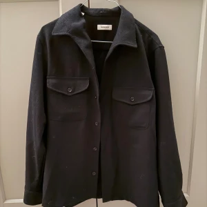 Mörkblå overshirt från Sandro - Stilren svart overshirt från Sandro med klassisk krage och två stora bröstfickor med lock och knapp. Rak passform och knäppning framtill. Perfekt lager-på-lager-plagg i ett mjukt, tjockare tyg som ger en avslappnad men snygg look.