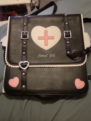 Svart ryggsäck med hjärtan och text - Säljer en svart ryggsäck i konstläder med vita och rosa detaljer. Framsidan har ett stort vitt hjärta med rosa kors och texten 'Sweet Girl', samt små rosa hjärtan och dekorativa nitar. Väskan har hjärtformade spännen och justerbara axelband. Perfekt för dig som gillar söt och unik stil.