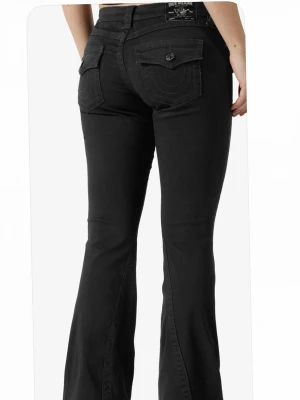 Svarta bootcut jeans True Religion - Snygga svarta bootcut jeans från True Religion med fickor bak som har lock och knapp. Jeansen har en klassisk femficksmodell och utsvängda ben. Perfekta för dig som gillar en tidlös men trendig look. Jeansen är bara använda 2 gånger så nästan helt nya!!