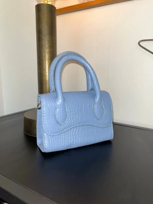 Ljusblå handväska med kedja - Säljer en ljusblå handväska i croco-mönstrad syntet med guldfärgad kedjerem, som går att ta av och på. Vet tyvärr inte vart den är ifrån💞