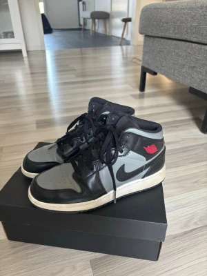Air Jordan 1 Mid svart/grå/röd - Snygga Air Jordan 1 Mid sneakers i svart och grått med röda detaljer. Klassisk siluett med svart Swoosh, perforerad tåbox och röda loggor på sidan och plösen. Ovandel i läder och textil, vita sulor och svarta skosnören. 