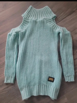 Mintgrön stickad tröja med cut-outs - Trendig mintgrön stickad tröja med grovstickat mönster, höga ribbade muddar och coola cut-outs vid axlarna. Hög krage och en dekorativ patch nertill. Perfekt för dig som vill sticka ut lite extra.