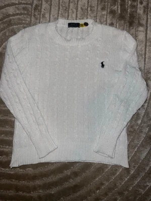 Vit kabelstickad tröja Polo Ralph Lauren - Säljer den för att den var för liten. Inga defekter hör av dig vid minsta lilla fråga!!                            Vit kabelstickad tröja från Polo Ralph Lauren med rund halsringning och klassisk broderad logga på bröstet. Tröjan har långa ärmar och ribbade muddar vid ärmslut och nederkant. Perfekt för en stilren och tidlös look.
