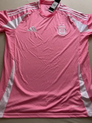 Rosa Argentina Adidas fotbollströja - Snygg rosa fotbollströja från Adidas med vita detaljer och AFA-logga på bröstet. Tröjan har korta ärmar, rund hals och är tillverkad i lätt och ventilerande material med Aeroready-teknologi. Perfekt för träning eller match.