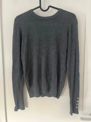 Mörkgrå rundhalsad tröja - En stilren mörkgrå tröja med rund halsringning och långa ärmar. Tröjan har dekorativa knappar i guld på ärmarna. Den är ifrån Zara och är i storlek S Använd ett få antal gånger 