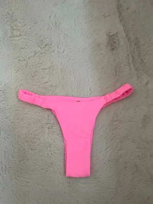 Rosa bikiniunderdel med rynkade band - Säljer en snygg rosa bikiniunderdel med rynkade band i sidorna. Tillverkad i stretchigt material som består av polyamid och elastan. Perfekt för strandhäng eller poolparty i sommar! Endast använd en gång för provning!  