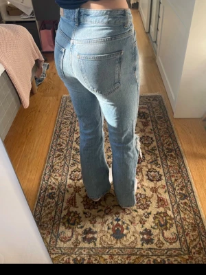 Ljusblå bootcut jeans med slitning - Säljer ett par ljusblå jeans med midwaist  Jeansen har en snygg slitning vid ena knät och klassiska femficksdetaljer. Perfekta för en avslappnad och trendig look.