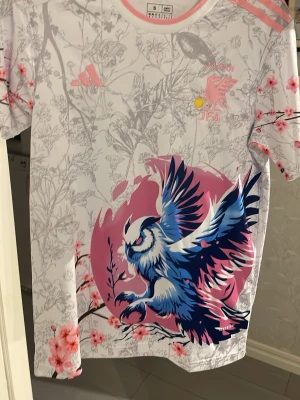Japan Adidas t-shirt med körsbärsblommor - Unik vit Adidas t-shirt för Japan med grått blommönster, rosa körsbärsblommor och en stor blå/vit fågel på framsidan. Rosa detaljer på axlarna och loggor. Perfekt för dig som gillar snygga och kreativa sporttröjor.