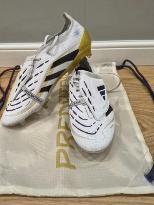 Adidas Predator fotbollsskor vit/guld - Snygga Adidas Predator fotbollsskor i vitt med svarta och lila detaljer samt guld på hälen och sulan. Skorna har klassisk Predator-design med täckt snörning och mönstrad ovandel för extra grepp på bollen. Levereras med originalpåse.