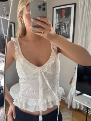 Zara Vit spetsig linne - Söt vit linnetopp med spetsdetaljer och volangkant. Toppen har breda axelband, v-ringning och knyt framtill. Perfekt för sommarens alla tillfällen och ger en romantisk vibe. Materialet är bomull med broderad spets.