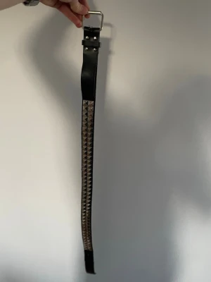 Svart bälte med silvernitar - Säljer ett svart bälte med silverfärgade pyramidnitar längs hela bandet. Bältet har ett klassiskt spänne i silver och är tillverkat i skinn. Perfekt för att ge din outfit en edgy touch.