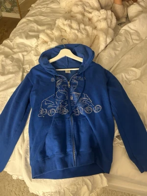 Ziphuddie med paljetter  - Blå hoodie med dragkedja framtill och huva. Framsidan har ett coolt motiv i strasstenar och text. Tillverkad i en mjuk bomullsblandning med fickor på magen. Perfekt för en avslappnad och trendig stil. Andvänd fåtal gånger köpt för 500kr