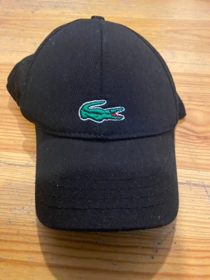 Svart keps från Lacoste - Snygg svart keps från Lacoste med klassisk böjd skärm och broderad grön krokodil-logga framtill. Kepsen är i bomull och har justerbar rem baktill för perfekt passform. Perfekt accessoar för en sportig och stilren look.
