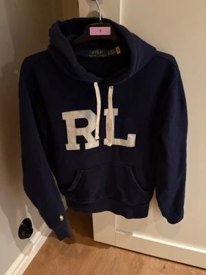 Mörkblå hoodie från Polo Ralph Lauren - Mörkblå hoodie från Polo Ralph Lauren med stora RL-bokstäver i vitt framtill, huva med vita dragsnören och känguruficka. Klassisk och stilren design med liten gul brodyr på ärmen. Perfekt för en avslappnad och trendig look.