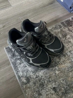 Svarta Asics - Säljer ett par svarta Asics sneakers med silvriga detaljer och meshpaneler för extra ventilation. Skorna har en chunky sula och klassisk snörning. Perfekta för dig som gillar sportig stil och vill ha bekväma skor till vardags.
