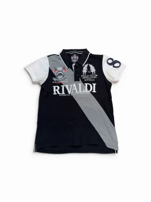 Svartvit Rivaldi t-shirt med tryck - Sportig svart och vit t-shirt från Rivaldi med grå diagonal rand och stora tryckdetaljer fram och bak. Kortärmad modell med krage och knappar, samt siffror på ärmarna. Tillverkad i bomull och har normal passform.