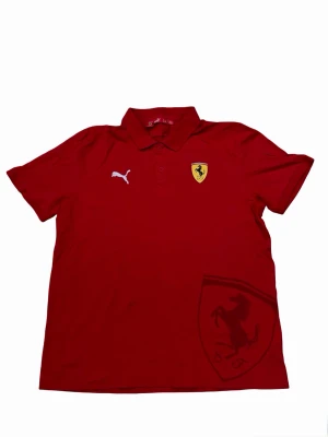 Röd Puma Ferrari t-shirt med krage - Röd t-shirt från Puma i samarbete med Ferrari. Klassisk pikékrage, korta ärmar och knappar framtill. Ferrari-logga på bröstet och stor hästgrafik nedtill. Tillverkad i mjuk bomull för skön känsla.