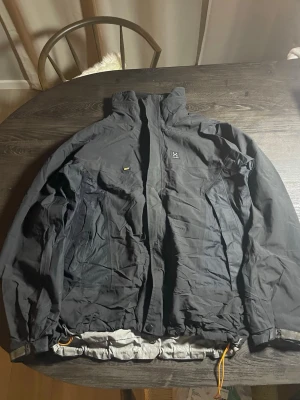 Svart vindjacka från Haglöfs - Säljer en svart vindjacka från Haglöfs med hög krage och dragkedja. Jackan har två sidofickor med dragkedja och justerbar nederkant med snören. Materialet är slitstarkt och jackan har Gore-Tex-teknologi för extra skydd mot vind och regn.