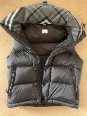 Svart dunväst med rutigt foder - Snygg svart dunväst från Burberry med stor huva och klassiskt rutigt foder i grått, vitt och svart. Västen har två dragkedjeförsedda fickor och stängs med både dragkedja och knappar. Perfekt för lager-på-lager och streetstyle.