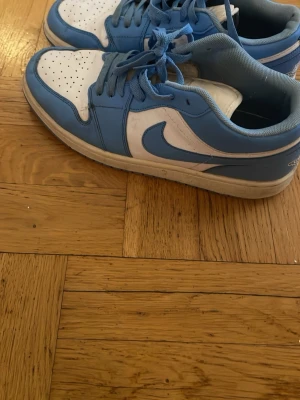 Nike Air Jordan 1 Low UNC blå/vit - Säljer ett par Nike Air Jordan 1 Low i UNC-färg, med ljusblått och vitt läder. Klassisk swoosh på sidan och perforerad tåbox för extra ventilation. Skön passform och ikonisk design som alltid är trendig.