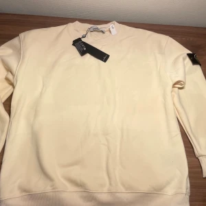 Stone Island beige hoodie - Tryck på bilderna för att se tydligare. Snygg beige hoodie från Stone Island med klassisk logotyp-patch på ärmen. Hoodien har huva med dragsko och ribbade muddar. Tillverkad i mjukt bomullsmaterial som känns skönt mot huden. Perfekt för dig som vill ha en stilren och trendig look. Storlek S. Riktigt mysig, skön och puffig. 