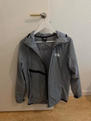Ljusgrå vindjacka från The North Face - Snygg ljusgrå vindjacka från The North Face med svart dragkedja och detaljer. Jackan har huva, två sidofickor och är tillverkad i ett lätt, vindtätt material. Perfekt för blåsiga dagar och enkel att matcha med olika outfits.