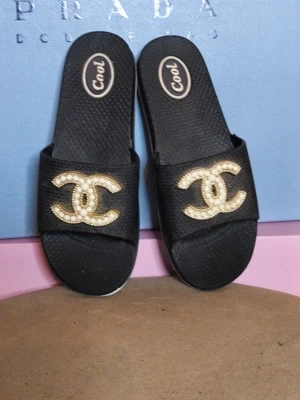 Svarta tofflor med pärldetaljer - The perfect mix of luxury-inspired style and everyday comfort. These black slides feature a stunning pearl and gold-tone logo detail that instantly elevates any summer outfit. ✨ Elegant pearl-embellished logo detail ✨ Lightweight and comfortable design ✨ Easy slip-on style ✨ Perfect for holidays, poolside, beach days or everyday wear ✨ Timeless black colour that matches everything Style them with linen trousers, denim shorts, a maxi dress or your favourite resortwear for an effortlessly chic loo