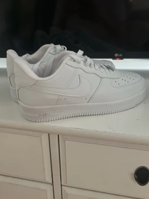 Nike Air Force 1 vita sneakers - Klassiska Nike Air Force 1 sneakers i helvitt läder med vit sula och vita skosnören. Modellen har låg profil, perforerad tåbox och ikonisk Swoosh-logga på sidorna. Perfekta för en clean och stilren look.