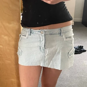 Ljusblå jeanskjol från S.M.L - Supersnygg ljusblå jeanskjol från S.M.L med coola stjärnpatchar och fickdetaljer. Kjolen är kort och har en klassisk femficksdesign med bälteshällor och dragkedja fram. 