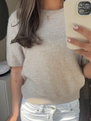 Beige stickad topp med kort ärm - Mysig beige stickad topp med rund hals och korta ärmar. Perfekt för lager på lager och enkel att matcha med olika stilar. Ribbad kant vid ärmslut och nederkant för en clean look.