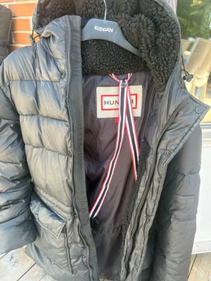 Svart dunjacka från Hunter - Snygg svart dunjacka från Hunter med huva och mjukt, fluffigt foder. Jackan har dragkedja, stora fickor och coola detaljer som randigt band på insidan. Perfekt för kalla vinterdagar och riktigt varm tack vare dunfyllningen.